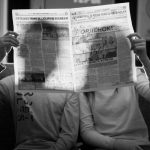A couple sitting indoors reading a newspaper in a black and white photograph.
