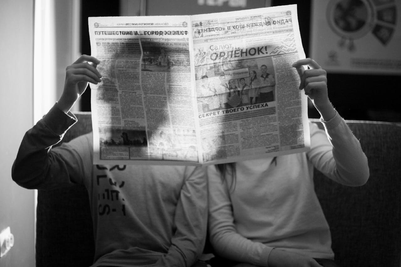 A couple sitting indoors reading a newspaper in a black and white photograph.